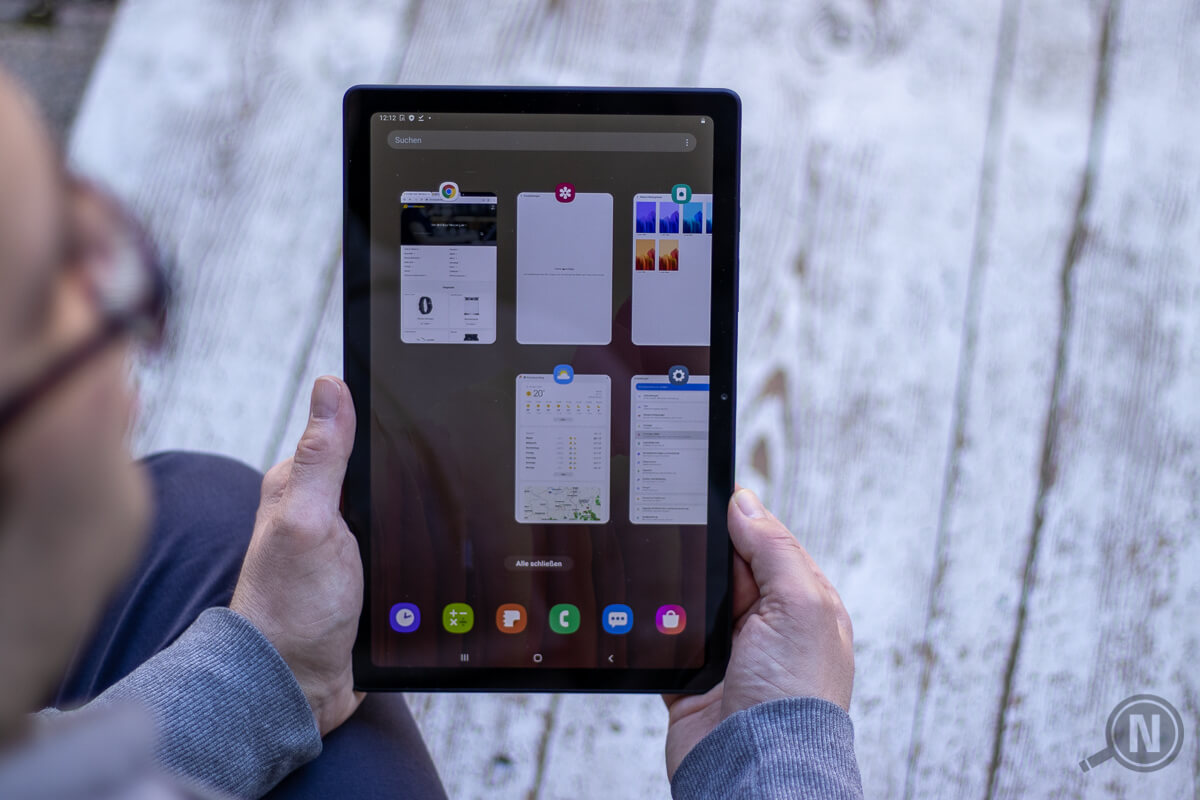 Blick über die rechte Schulter eines Mannes, der ein Tablet mit beiden Händen hält. Gesicht und Brille sind unscharf am linken Bildrand zu erkennen, im Hintergrund einen weiß gemaserte Holzplatte.