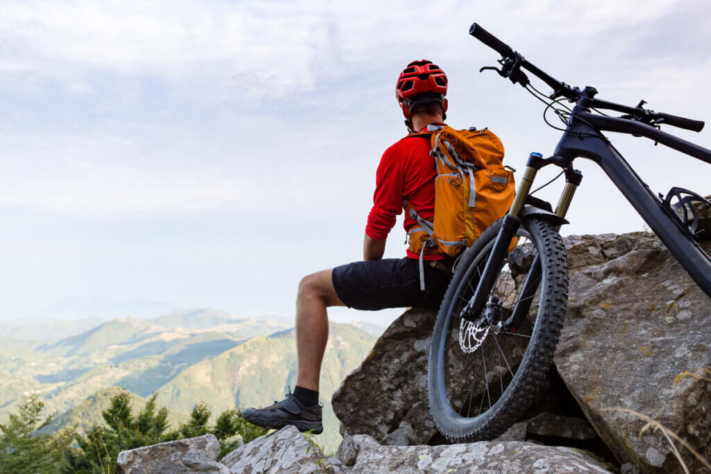 radfahrer sitzt mit rucksack auf einem fels und schaut sich die berglandschaft an