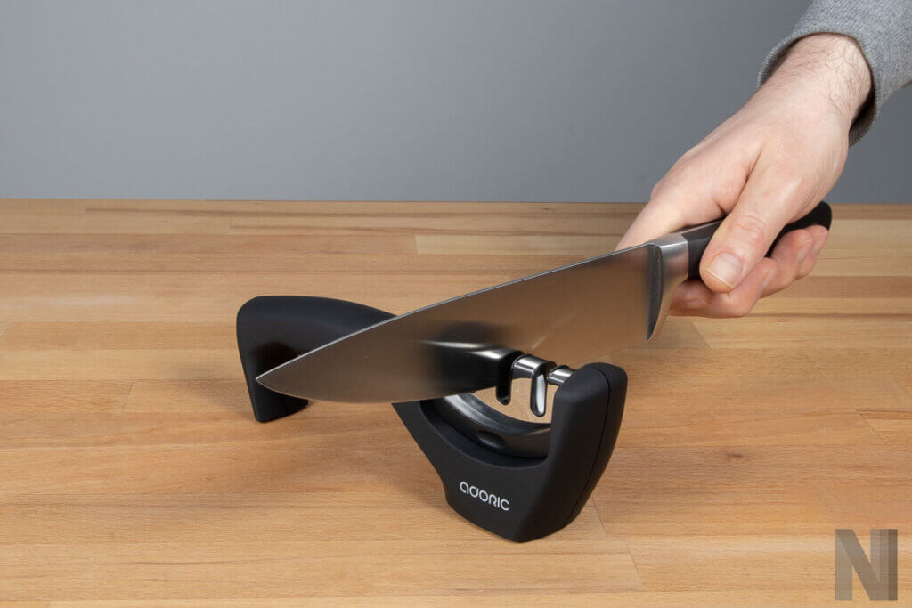 Durchziehschleifer mit Handgriff auf einer hellen Tischplatte, Hand zieht großes Messer durch einer der Schlitze des Messerschaerfers.