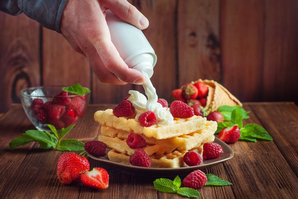 Person sprueht Sahne auf Waffeln mit Himbeeren