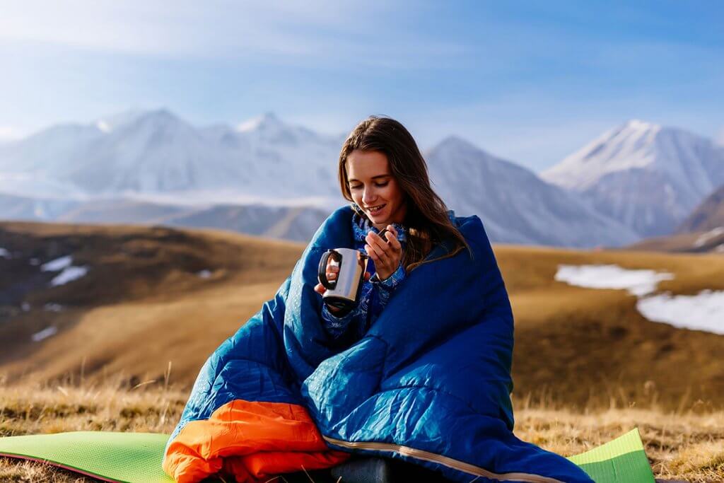 Frau sitzt in Berglandschaft in Schlafsack gehuellt und trinkt ein Heissgetraenk